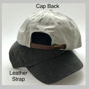May include: A gray and black baseball cap with a leather strap and a metal buckle. The back of the cap says "Cap Back" and the strap says "Leather Strap".