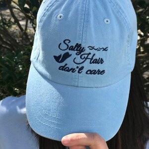 May include: A light blue baseball cap with the text "Salty Hair don't care" embroidered in black. A small bird graphic is also embroidered on the cap.