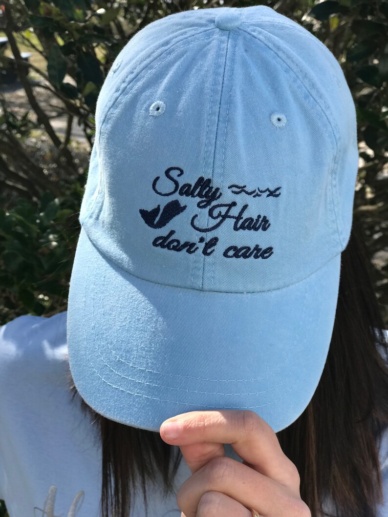 May include: A light blue baseball cap with the text "Salty Hair don't care" embroidered in black. A small bird graphic is also embroidered on the cap.