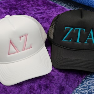 Könnte beinhalten: Zwei Trucker-Caps, eine weiße mit rosa Stickerei, die "ΔZ" sagt, und eine schwarze mit blauer Stickerei, die "ZTA" sagt.