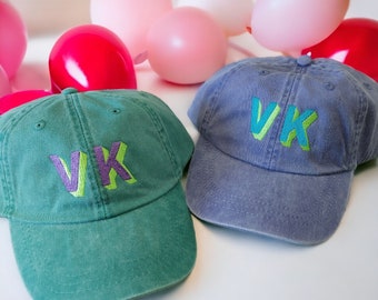 Könnte beinhalten: Zwei Baseballkappen, eine grüne und eine blaue, mit den Buchstaben "VK" in Grün, Lila und Blau gestickt.