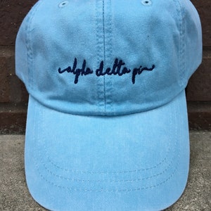 Puede incluir: Una gorra de béisbol azul claro con el texto "alpha delta pi" bordado en negro en la parte delantera.