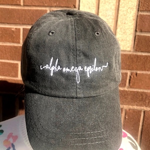Puede incluir: Gorra de béisbol negra con el texto bordado "alpha omega epsilon" en escritura blanca. La gorra tiene una visera curva y un cierre de correa de tela. El sombrero está hecho de un material suave y desgastado.