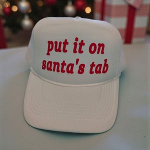 Könnte beinhalten: Weißer Trucker-Hut mit dem Schriftzug "put it on santa's tab" in roter Schrift. Der Hut hat eine weiße Frontplatte und eine weiße Mesh-Rückseite. Ein lustiges, festliches Accessoire für die Weihnachtszeit.