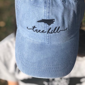 Pode incluir: Um bon&eacute; de beisebol de denim azul claro com um contorno bordado do estado da Carolina do Norte e o texto "tree hill" em preto.