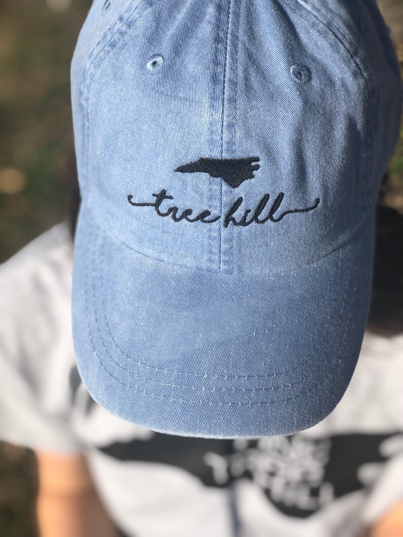 Pode incluir: Um bon&eacute; de beisebol de denim azul claro com um contorno bordado do estado da Carolina do Norte e o texto "tree hill" em preto.