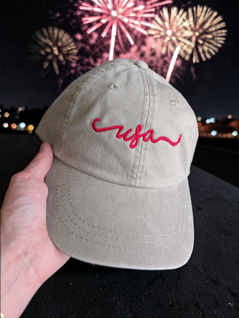 May include: A khaki baseball cap with the red embroidered word "usa". The cap is held up against a night sky with fireworks in the background. The cap has a curved brim and a rounded crown.