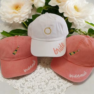 May include: Three baseball caps, one white and two coral pink, with embroidered text. The white cap says "bride" and has a gold ring design. The coral pink caps say "bridesmaid" and have a champagne bottle design.