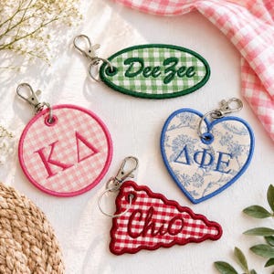 May include: Four embroidered keychains with metal lobster clasps. One oval green gingham with 'DeeZee', one pink gingham circle with 'KA', one heart-shaped with blue floral print and 'ΔΦΕ', and one red gingham triangle with 'ChiO' embroidered text.
