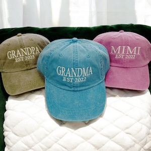 May include: Three baseball caps with embroidered text. The caps are green, blue, and pink. The text on the caps reads "Grandpa Est 2025", "Grandma Est 2022", and "Mimi Est 2022".