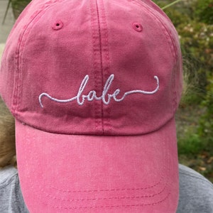 May include: A pink baseball cap with the word "babe" embroidered in white script across the front. The cap has a curved brim and a fabric strap closure. The hat is made of a soft, textured material.