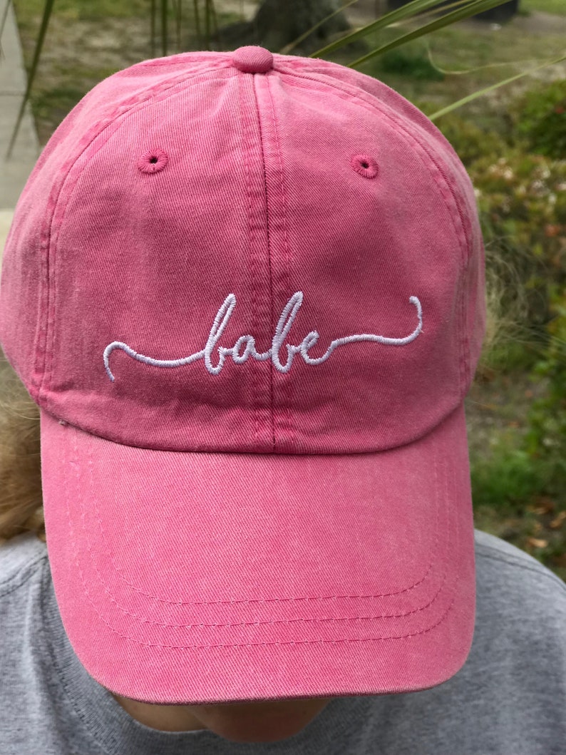 May include: A pink baseball cap with the word "babe" embroidered in white script across the front. The cap has a curved brim and a fabric strap closure. The hat is made of a soft, textured material.