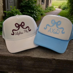 Puede incluir: Dos gorras de camionero con la parte trasera de malla blanca y viseras curvas. Una gorra es blanca con un lazo granate y el texto "Alpha Phi". La otra es azul claro con un lazo azul claro y el texto "AXiD".
