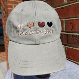 May include: A light green baseball cap with an embroidered message that reads "love everybody". The message is in a white font and is accompanied by four embroidered hearts in different skin tones.