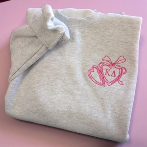 Puede incluir: Una sudadera gris claro con cuello interior y puños blancos. La sudadera presenta un diseño bordado en rosa de dos corazones, un lazo y las letras "KD". La sudadera está doblada sobre una superficie rosa.