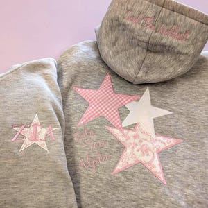 Peut inclure: Un sweat à capuche gris avec des appliqués d'étoiles roses. Une étoile a un motif vichy, une autre un motif floral. La capuche porte la broderie "Seek The Noblest". La manche gauche porte une étoile avec "ZTA" brodé.