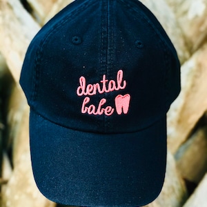 May include: A black baseball cap with a pink embroidered design that says "dental babe" with a tooth graphic.