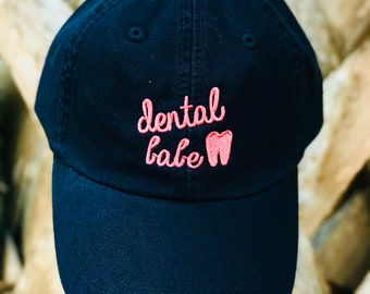May include: A black baseball cap with a pink embroidered design that says "dental babe" with a tooth graphic.