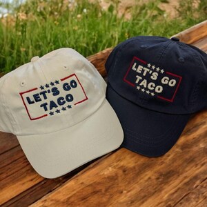May include: Two baseball caps, one cream and one navy blue, are displayed. Both caps feature the embroidered text "LET'S GO TACO" within a red and blue rectangular border, with stars above and below the text. The caps are on a wooden surface.