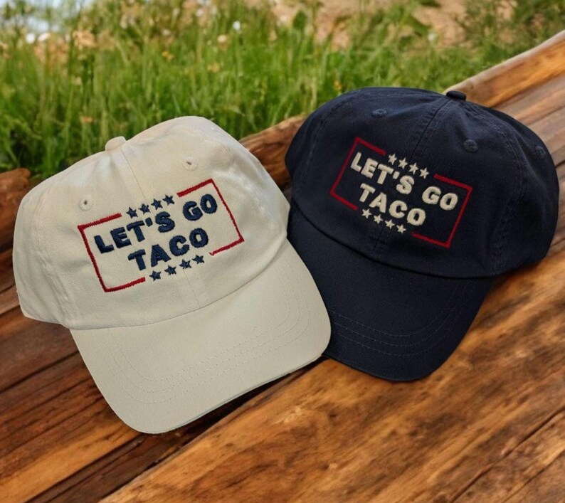 May include: Two baseball caps, one cream and one navy blue, are displayed. Both caps feature the embroidered text "LET'S GO TACO" within a red and blue rectangular border, with stars above and below the text. The caps are on a wooden surface.