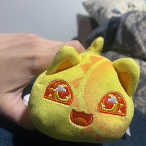 May include: A plush toy with a yellow body and orange accents. The toy has embroidered eyes with a sun and cloud design, small ears, and a smiling mouth. The toy is held in a person's hand.