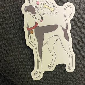 Könnte beinhalten: Ein Aufkleber mit einem Cartoon-Hund in Grau und Weiß mit einem roten Halsband und einem Knochen in seiner Gedankenblase. Der Hund schaut mit einem fragenden Ausdruck nach oben und seine Zunge hängt heraus.