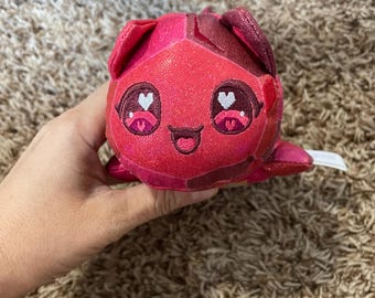 May include: A small, plush toy shaped like a faceted crystal or gem in shades of vibrant red and magenta. The surface has a subtle glittery texture. It features embroidered eyes with white heart-shaped highlights and a small, smiling mouth.