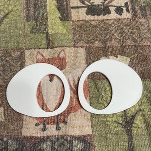 May include: Two white, oval-shaped objects with a circular cutout in the center. The objects are placed on a patterned fabric with a fox and owl design. The fabric has a mix of green, brown, and beige colors.