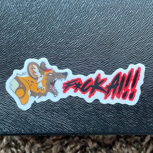 May include: A sticker featuring a cartoon African wild dog with an open mouth and the text "F*CK ALL!!!" in red and black. The sticker is on a textured black surface.
