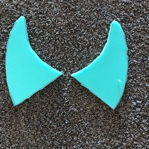 以下が含まれることがあります： 三日月型のターコイズブルーのサーフボードフィン2枚が、茶色のカーペットの上に置かれています