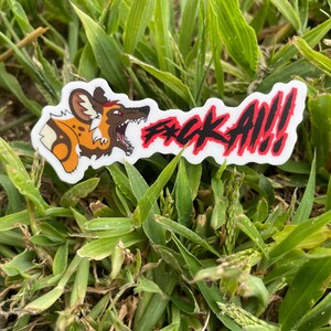 Könnte beinhalten: Ein weißer Aufkleber mit einer Cartoon-Illustration eines afrikanischen Wildhundes und dem Text "F*CK ALL!!" in Rot und Schwarz. Der Aufkleber liegt auf grünem Gras.