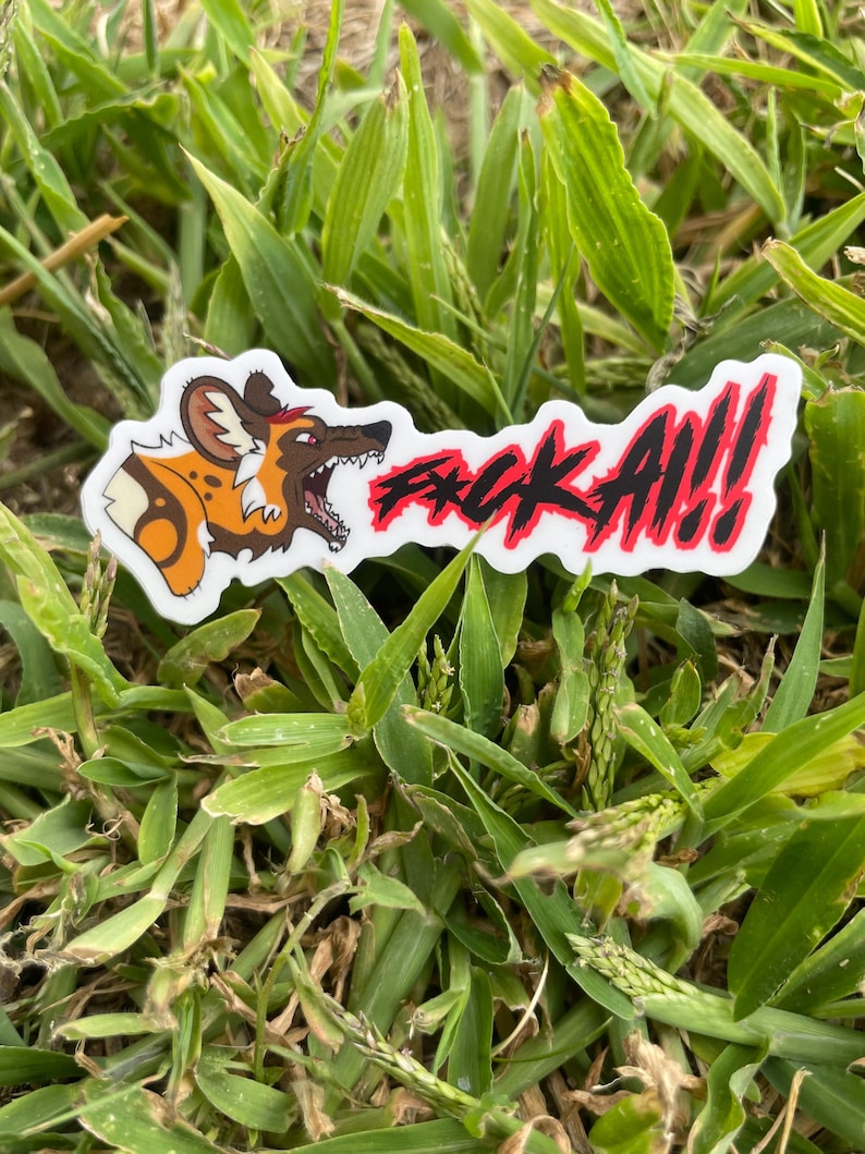 May include: A white sticker featuring a cartoon African wild dog illustration and the text "F*CK ALL!!" in red and black. The sticker is placed on green grass.