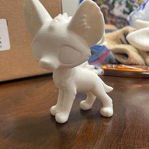 以下が含まれることがあります： 大きな耳を持つスタイリッシュな犬の白い3Dプリントフィギュア。フィギュアは木製の表面に立っています