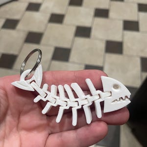 May include: A white, 3D-printed fish skeleton keychain. The keychain features a detailed design of a fish skeleton with articulated joints and a metal keyring for attaching to keys or bags.
