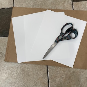 May include: A pair of silver scissors with a patterned handle rests on a stack of white paper. The scissors are open, and the blades are sharp. The paper is on top of a brown cardboard surface.