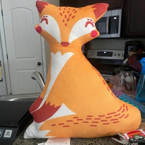 May include: Orange and red fox-shaped plush pillow with white accents. The pillow has a simple, stylized design with a white belly and red accents around the eyes and ears. The pillow is sitting on a table with a tag attached that reads "Made for: Triangle Home Fashions 120 Tices Lane, East Brunswick, NJ 08816".