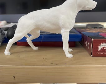 May include: A white, 3D-printed figurine of a dog in a standing pose. The dog has a muscular build and a short, smooth coat. The figurine is placed on a wooden shelf, with other items in the background.