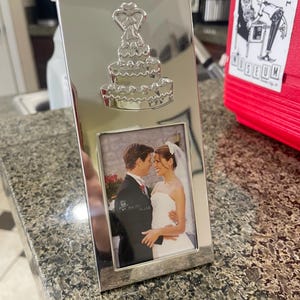 May include: A silver-colored photo frame with a wedding cake design at the top. The frame holds a photo of a couple in wedding attire. The frame is rectangular and sits on a speckled countertop.