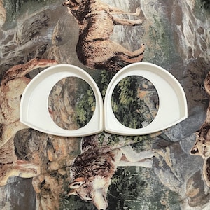 May include: Two white plastic eye-shaped frames, possibly for a costume or craft project. The frames are on a fabric background with a wolf print.