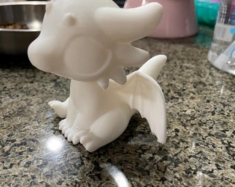 May include: A white 3D printed dragon figurine with a wide wingspan and large eyes. The dragon is sitting on a worktop.