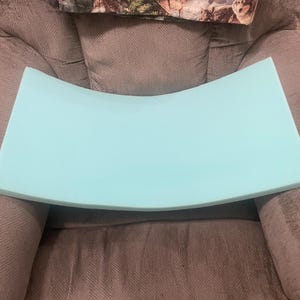 May include: A light blue, curved foam cushion. The cushion is rectangular with a gentle arch. The background is a grey upholstered chair with a wolf-themed pillow.