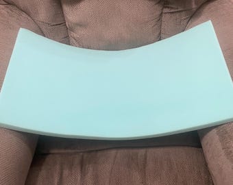May include: A light blue, curved foam cushion. The cushion is rectangular with a gentle arch. The background is a gray upholstered chair with a wolf-themed pillow.
