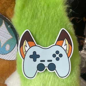 Op de afbeelding: Een sticker met een lichtblauwe videogamecontroller met donkerblauwe knoppen en joysticks. De controller is voorzien van twee puntige dierenoren die aan de buitenkant bruin zijn en aan de binnenkant oranje met witte accenten.