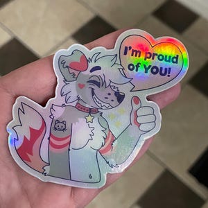May include: A holographic sticker featuring a cartoon wolf with a red and white striped arm, a star on its collar, and a cat tattoo. The wolf is giving a thumbs up and a heart-shaped speech bubble says "I'm proud of you!"
