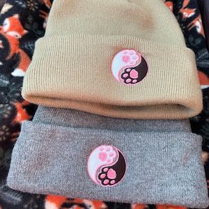 May include: Two knit beanies with a yin yang symbol featuring pink paw prints. The top beanie is beige and the bottom beanie is grey.