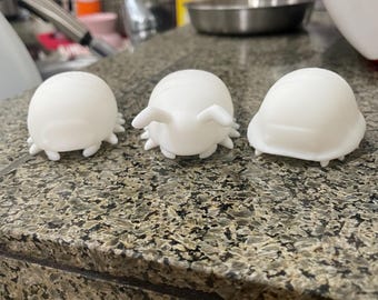 May include: Three white, insect-shaped figurines with rounded bodies and small legs are arranged on a speckled countertop. The figurines have a smooth, matte finish and are positioned in a row, showcasing their unique design.