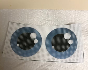 May include: Two cartoon eye decals. Each eye features a light blue iris, a black pupil, and white highlights. The decals are on white, textured paper.