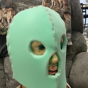 May include: A light green foam mask with eye and mouth openings. The mask is sewn together with a grey thread.
