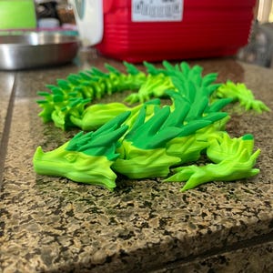 May include: A segmented, articulated dragon figurine in shades of green. The dragon's head and body are a bright, lime green, with darker green accents along the spine and wings. The dragon is posed on a speckled countertop.