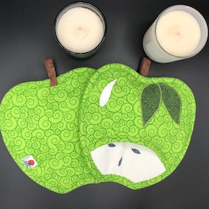 May include: A green apple-shaped pot holder with a swirl pattern, white accents, and brown fabric loops. Two candles in glass jars are in the background. The pot holder is made of fabric.